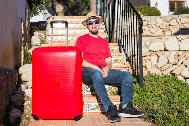 Concetto di viaggio, turismo e persone - uomo felice che si siede sulle scale in un cappello con la valigia rossa.