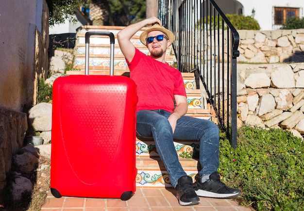 Concetto di viaggio, turismo e persone - uomo felice che si siede sulle scale in un cappello con la valigia rossa.