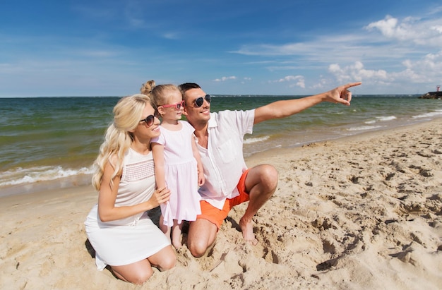 concetto di viaggio, vacanza, adozione e persone - famiglia felice con una bambina in occhiali da sole che punta il dito sulla spiaggia estiva