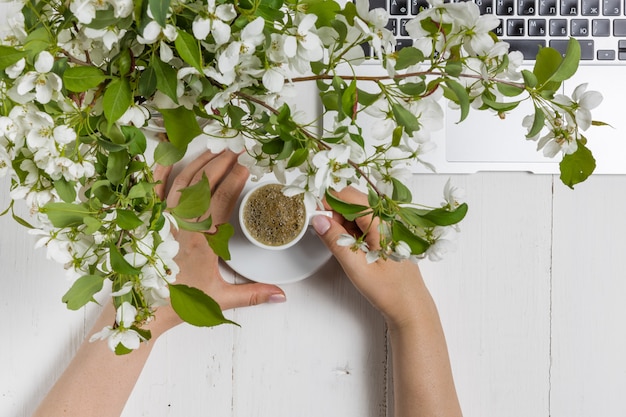 Concetto femminile di affari con il computer portatile, il mazzo dei fiori della molla e le mani della donna che tengono tazza di tè o caffè caldo