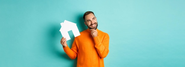 Concetto immobiliare bell'uomo adulto in maglione arancione che pensa all'acquisto di un appartamento o al noleggio di un holdi