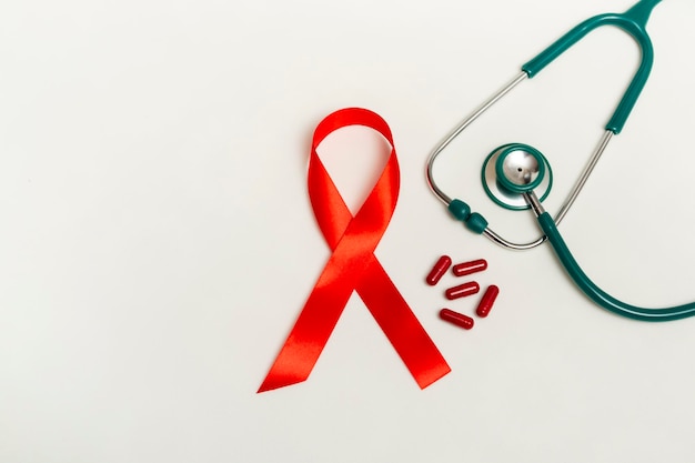 Concetto medico per la Giornata mondiale contro l'AIDS di dicembre. Nastro rosso, stetoscopio medico verde e pillole o vitamine rosse di terapia su un fondo bianco. Chiudere lo spazio della copia