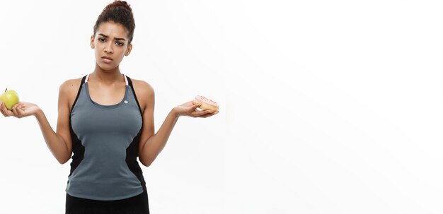 Concetto sano e dietetico Il bellissimo afroamericano sportivo prende una decisione tra la ciambella e la mela verde isolata su sfondo bianco