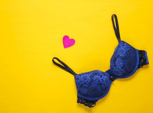 Concetto sessuale romantico. Bellissimo reggiseno sexy, cuore decorativo su un tavolo giallo. Vista dall'alto, minimalismo