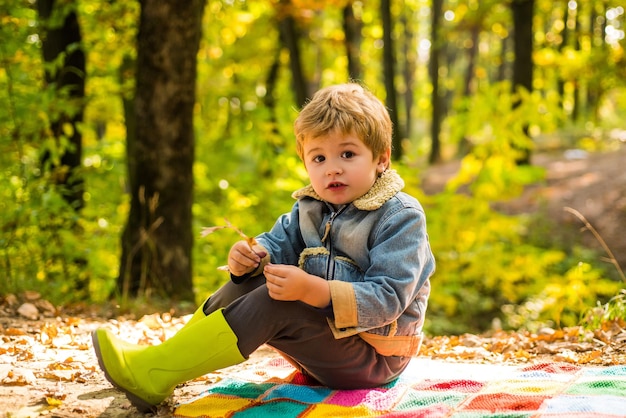 Concetto turistico carino. Gli piace fare escursioni. Il bambino si siede sul picnic della foresta a quadri. Il bambino si rilassa nella foresta autunnale. La scuola forestale è educazione all'aria aperta. Visita gli spazi naturali. Ragazzo in stivali di gomma che si rilassa nella foresta