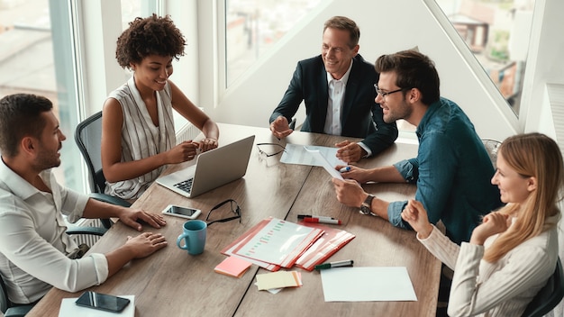 Condividere nuove idee. Gruppo di giovani uomini d'affari che discutono di qualcosa e sorridono seduti al tavolo dell'ufficio. Brainstorming.