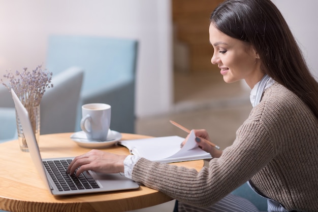 Condizioni confortevoli. Felice giovane donna graziosa che sorride e che utilizza computer portatile mentre prende appunti.