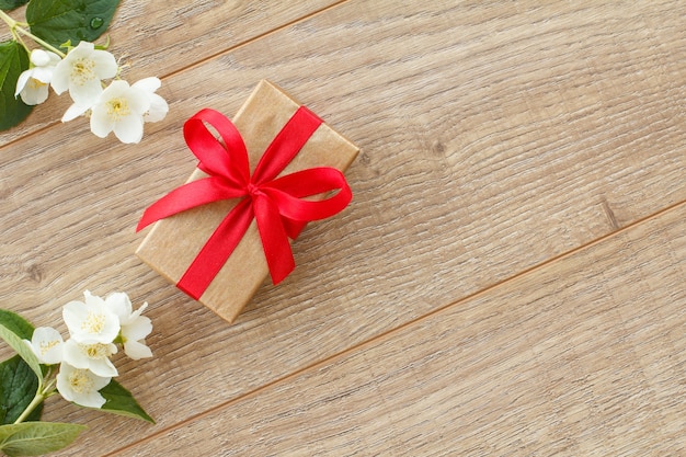 Confezione regalo avvolta con nastro rosso su tavole di legno decorate con fiori di gelsomino. Vista dall'alto con copia spazio.