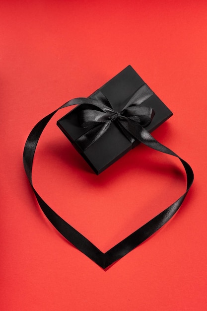 Confezione regalo avvolta in nero con fiocco nero e nastro disposto come cuore su rosso Spazio di copia Ripresa verticale