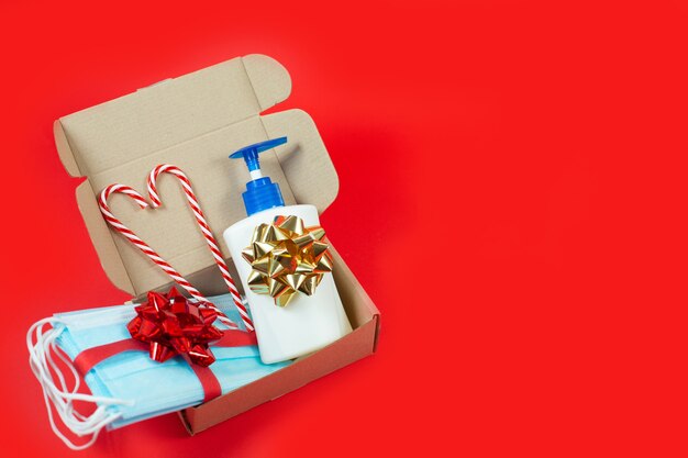 Confezione regalo di Natale con maschere usa e getta, disinfettante per le mani e bastoncino di zucchero su sfondo rosso.