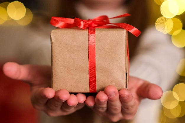 Confezione regalo di Natale nelle mani dei bambini. Piccola scatola regalo con nastro rosso su sfondo di luci scintillanti