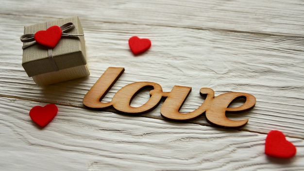 Confezione regalo, piccoli cuori rossi e scritta decorativa "Love" in legno su una scrivania bianca. Vista laterale. San Valentino sullo sfondo.