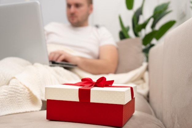 Confezione regalo rossa sullo sfondo dell'uomo sdraiato sul divano, al lavoro, navigando in internet al laptop, sotto il plaid al chiuso. Natale, San Valentino, concetto di shopping online per le vacanze