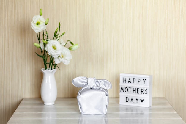 Confezione regalo trendy avvolta in tessuto di seta con tecnica Furoshiki, light box con scritta Happy Mother's Day, fiori bianchi Eustoma o Lisianthus in vaso su tavola di legno. Zero concetto di vita dei rifiuti.