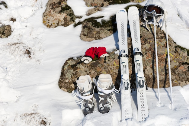 Configurazione degli sport invernali con guanti rossi, paio di sci, scarponi, casco e bastoncini appoggiati su una superficie di pietra parzialmente ricoperta di neve