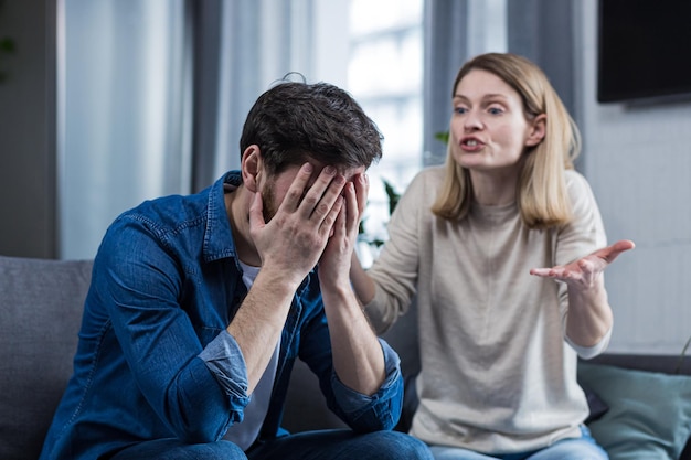 Conflitto familiare Litigio incomprensione La donna grida disperata al marito piangendo richiede spiegazioni L'uomo ascolta coprendosi il viso con le mani Seduto sul divano di casa