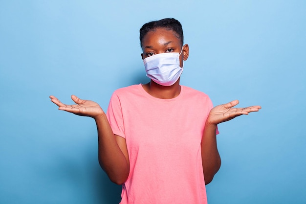 Confondere l'adolescente afroamericano con la maschera protettiva contro il coronavirus alzando le spalle guardando la telecamera in posa su sfondo blu in studio. Gesto di domanda incerta