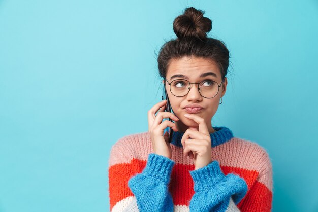 confusa bella donna in occhiali parlando su smartphone isolato su muro blu