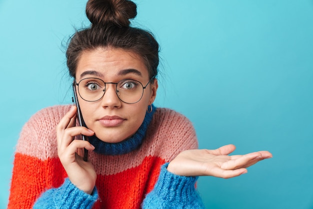 confusa bella donna in occhiali parlando su smartphone isolato su muro blu