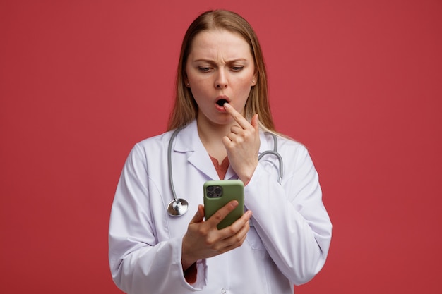 Confuso giovane dottoressa bionda che indossa abito medico e stetoscopio intorno al collo tenendo e guardando il telefono cellulare mantenendo il dito sul labbro