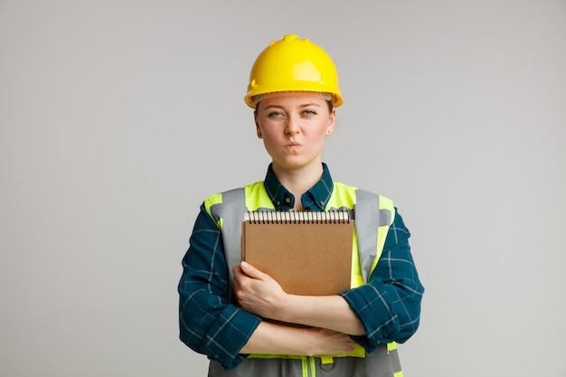 Confuso giovane operaio edile femminile che indossa il casco di sicurezza e giubbotto di sicurezza che abbraccia il blocco note con le labbra increspate