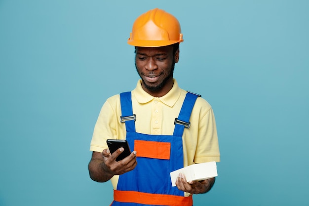 Confuso tenendo il mattone e guardando il telefono in mano giovane costruttore afroamericano in uniforme isolato su sfondo blu