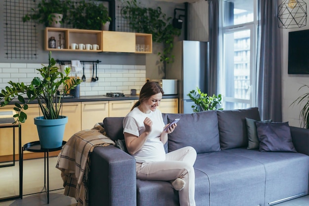 Congedo di maternità Bella donna incinta seduta sul divano a casa con in mano un telefono cellulare digitando messaggi chiamando guardando ricevere buone notizie Sorridere felice