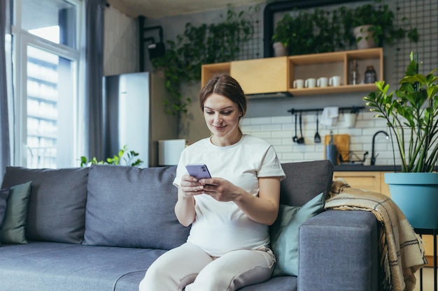 Congedo di maternità Bella donna incinta seduta sul divano di casa con in mano un telefono cellulare corrispondente nelle chat dei genitori digitando messaggi chiamando guardando ricevere buone notizie Sorridere felice