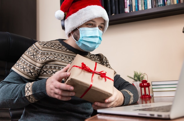 Congratulazioni in linea di Natale. Il giovane in cappello di Babbo Natale e maschera per il viso fa un regalo e parla utilizzando il laptop per videochiamare amici e bambini. Natale durante il coronavirus.