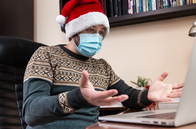 Congratulazioni in linea di Natale. Scioccato e sorpreso Il giovane con il cappello di Babbo Natale e la maschera per il viso parla utilizzando il laptop per videochiamare amici e bambini. Natale durante il coronavirus.