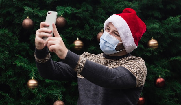 Congratulazioni online di Natale. Ritratto di uomo che indossa santa cappello e mascherina medica parlando al telefono con amici e genitori. bokeh albero di natale sullo sfondo