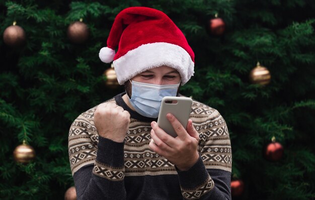 Congratulazioni online di Natale. Uomo allegro che porta il cappello della Santa e la mascherina medica parlando al telefono con amici e genitori. bokeh albero di natale sullo sfondo