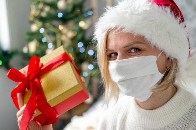 Congratulazioni per la maschera di Natale. Donna del ritratto nella mascherina medica e Santa con la confezione regalo