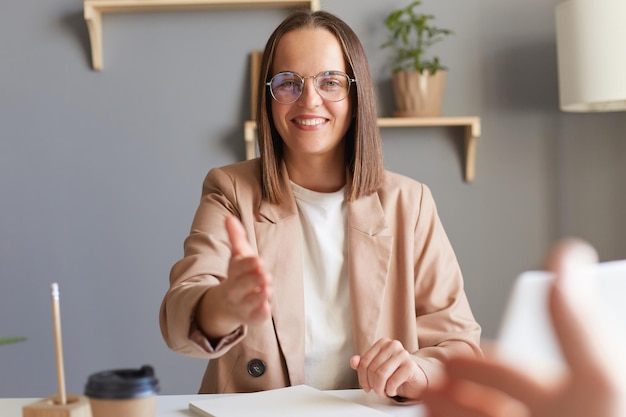 Congratulazioni sei stata assunta Inquadratura in interni di una donna dai capelli castani che indossa una giacca beige e occhiali seduti al suo posto di lavoro durante una riunione di lavoro con un saluto di stretta di mano