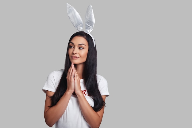 Coniglietto premuroso. Attraente giovane donna con orecchie da coniglio che guarda lontano e sorride mentre si trova su uno sfondo grigio