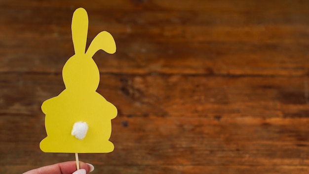 Coniglio giallo fatto di carta su uno spazzolino da denti. La mano tiene la decorazione di cottura di Pasqua. Biglietto di Pasqua su fondo in legno