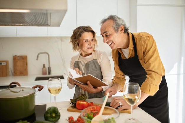 Coniugi anziani felici che controllano la ricetta nel libro di cucina mentre preparano cibo sano insieme in cucina