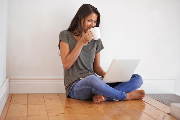 Connettività wireless Inquadratura di una giovane donna seduta per terra a casa che beve un caffè e usa un laptop
