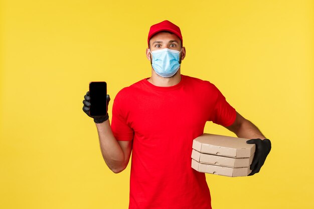 Consegna del cibo, monitoraggio degli ordini, covid-19 e concetto di auto-quarantena. Corriere entusiasta in uniforme rossa, maschera medica e guanti, che mostra l'app per ordinare la pizza, traccia l'applicazione su smartphone