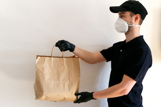 Consegna di alimenti e merci durante la pandemia di coronavirus. Fattorino in uniforme. Il fattorino tiene in mano un sacchetto di carta artigianale. Uomo in maschera protettiva medica e guanti di gomma.