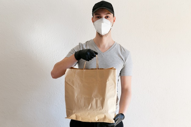 Consegna di alimenti e merci durante la pandemia di coronavirus. Fattorino in uniforme. Il fattorino tiene in mano un sacchetto di carta artigianale. Uomo in maschera protettiva medica e guanti di gomma.