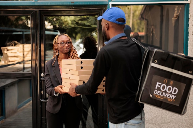 Consegna di cibo in ufficio, cliente allegro che tiene pile di scatole per pizza, donna che riceve l'ordine dal corriere all'aperto. Impiegato afroamericano che prende la pila di pacchetti pasto, uomo che consegna il pranzo