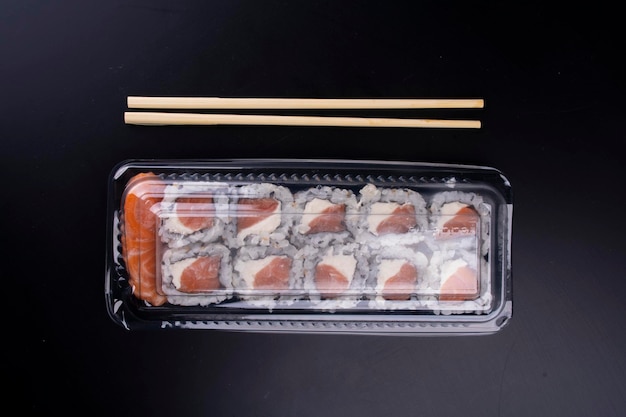 Consegna di cibo orientale giapponese con imballaggi in plastica chiusi e suhsis di salmone crudo con sashimi di formaggio cremoso e bacchette su sfondo nero vista dall'alto