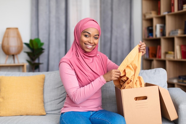 Consegna e shopping online felice signora afroamericana che tiene un pezzo di vestiti che disimballa