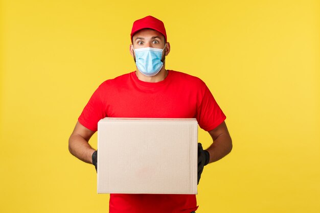 Consegna espressa durante la pandemia, covid-19, spedizione sicura, concetto di shopping online. Corriere sorpreso e scioccato in uniforme rossa, maschera medica e guanti, con in mano il pacco si sente stupito