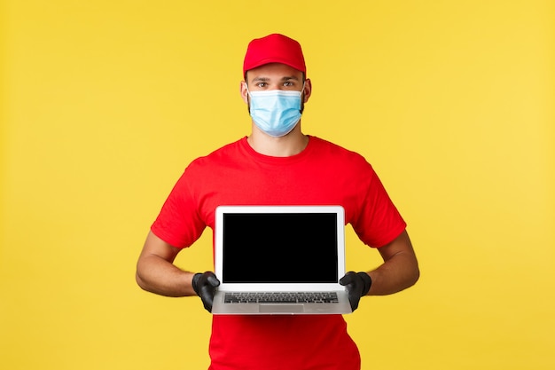 Consegna espressa durante la pandemia, covid-19, spedizione sicura, concetto di shopping online. Dipendente con berretto e guanti uniformi rossi, corriere in maschera medica che mostra il sito Web sullo schermo del laptop