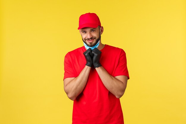 Consegna espressa durante la pandemia, covid-19, spedizione sicura, concetto di shopping online. Simpatico corriere barbuto con uno sguardo adorabile, maschera medica da decollo e macchina fotografica sorridente amichevole, indossa guanti