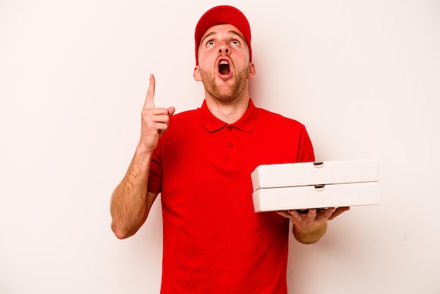 Consegna giovane uomo caucasico che tiene pizze isolate su sfondo bianco che punta verso l'alto con la bocca aperta