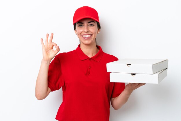 Consegna pizza donna caucasica con uniforme da lavoro che raccoglie scatole di pizza isolate su sfondo bianco che mostra il segno ok con le dita