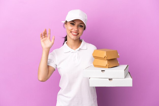 Consegna pizza Ragazza russa con divisa da lavoro che prende scatole per pizza e hamburger isolati su sfondo viola salutando con la mano con espressione felice